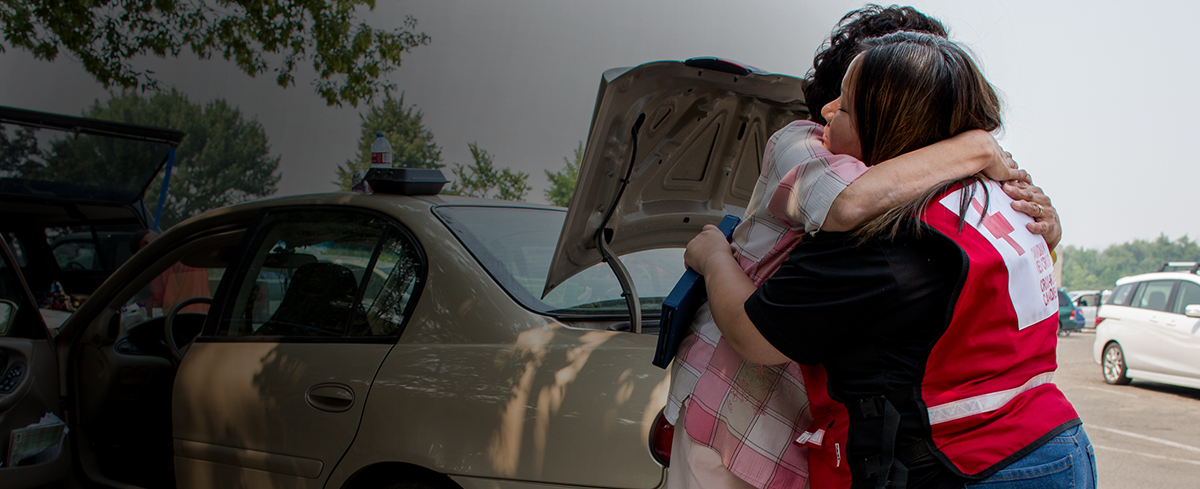 Une femme en chemise de la Croix-Rouge étreint une autre femme devant une voiture, témoignant de la compassion.