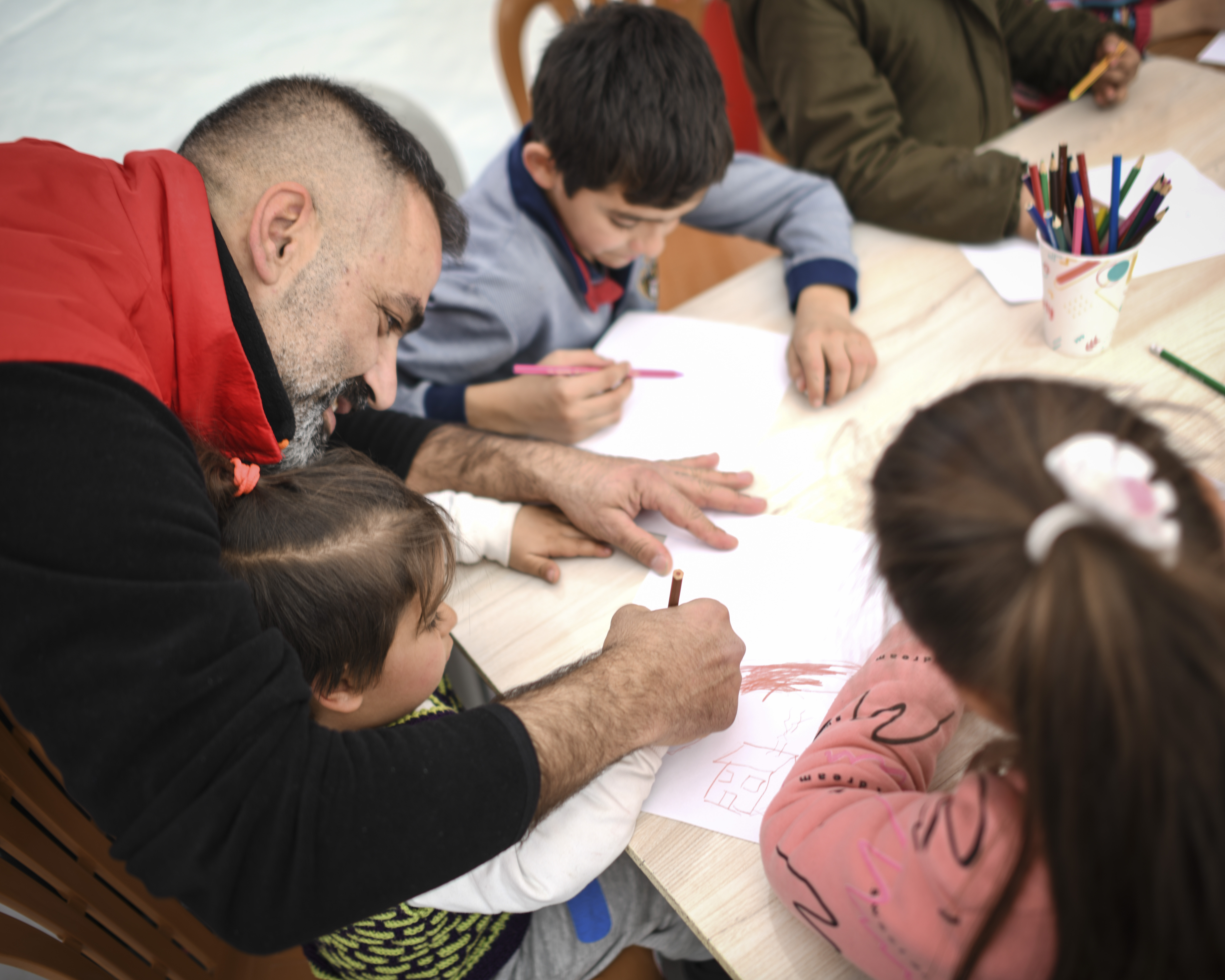 Trois enfants assis à une table dessinent sous l’œil d’un bénévole en dossard rouge.