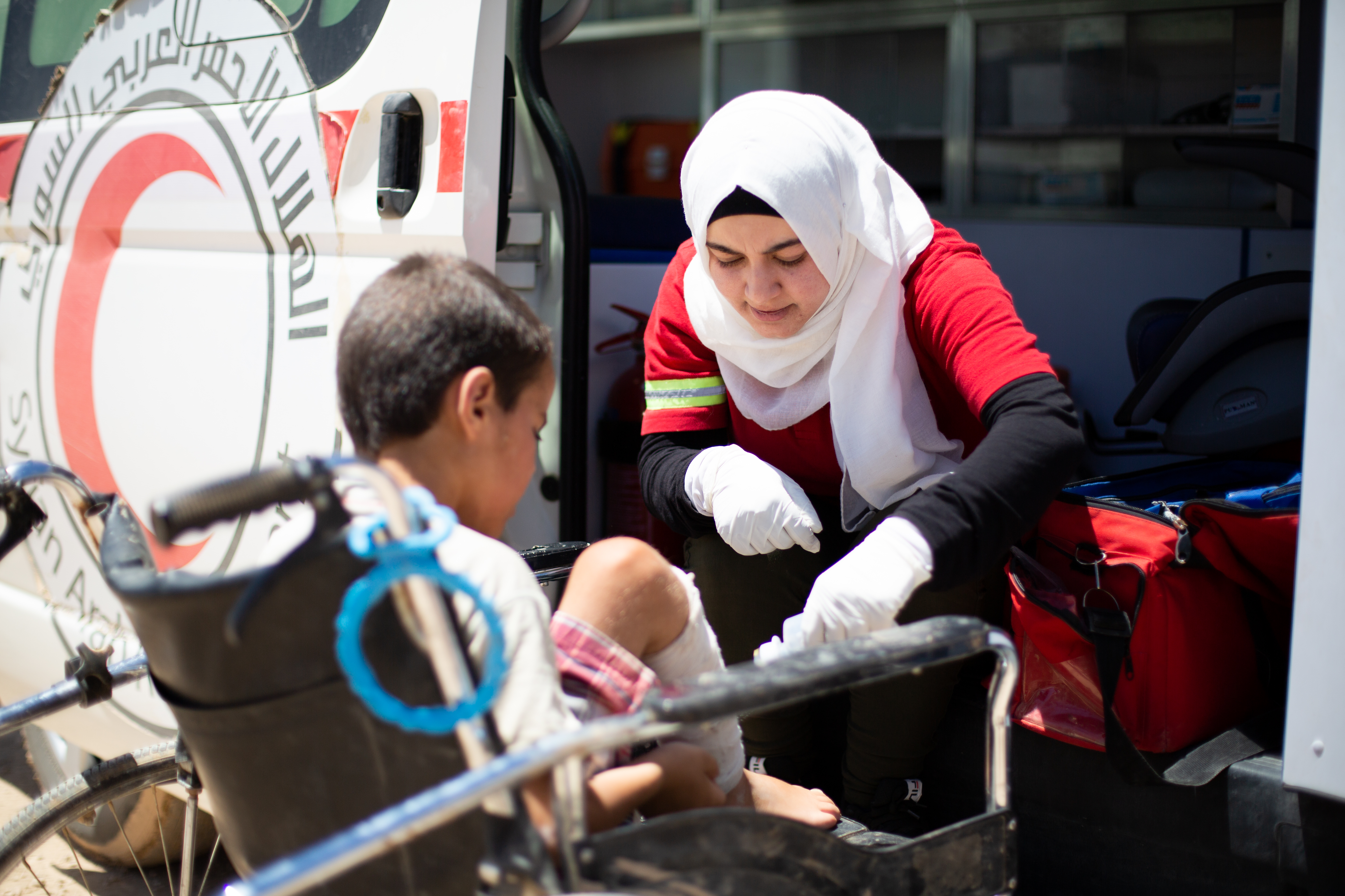 Une femme portant un foulard blanc sur sa tête et un uniforme rouge administre des soins à un enfant en fauteuil roulant.