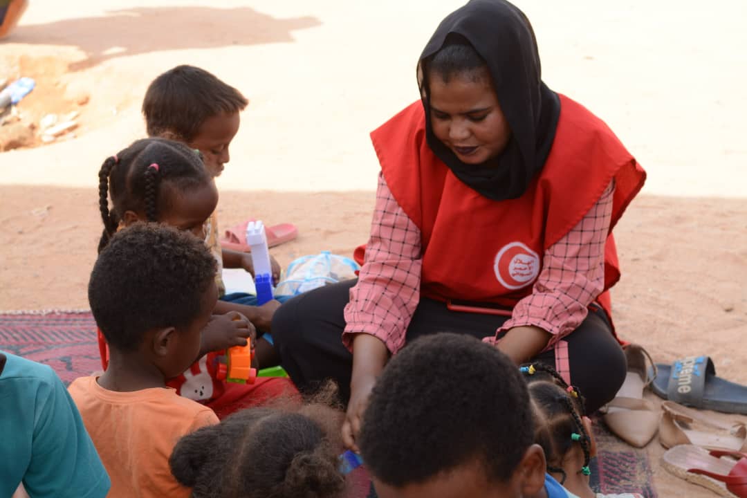 Une femme portant un dossard rouge est assise dehors par terre et joue avec des enfants