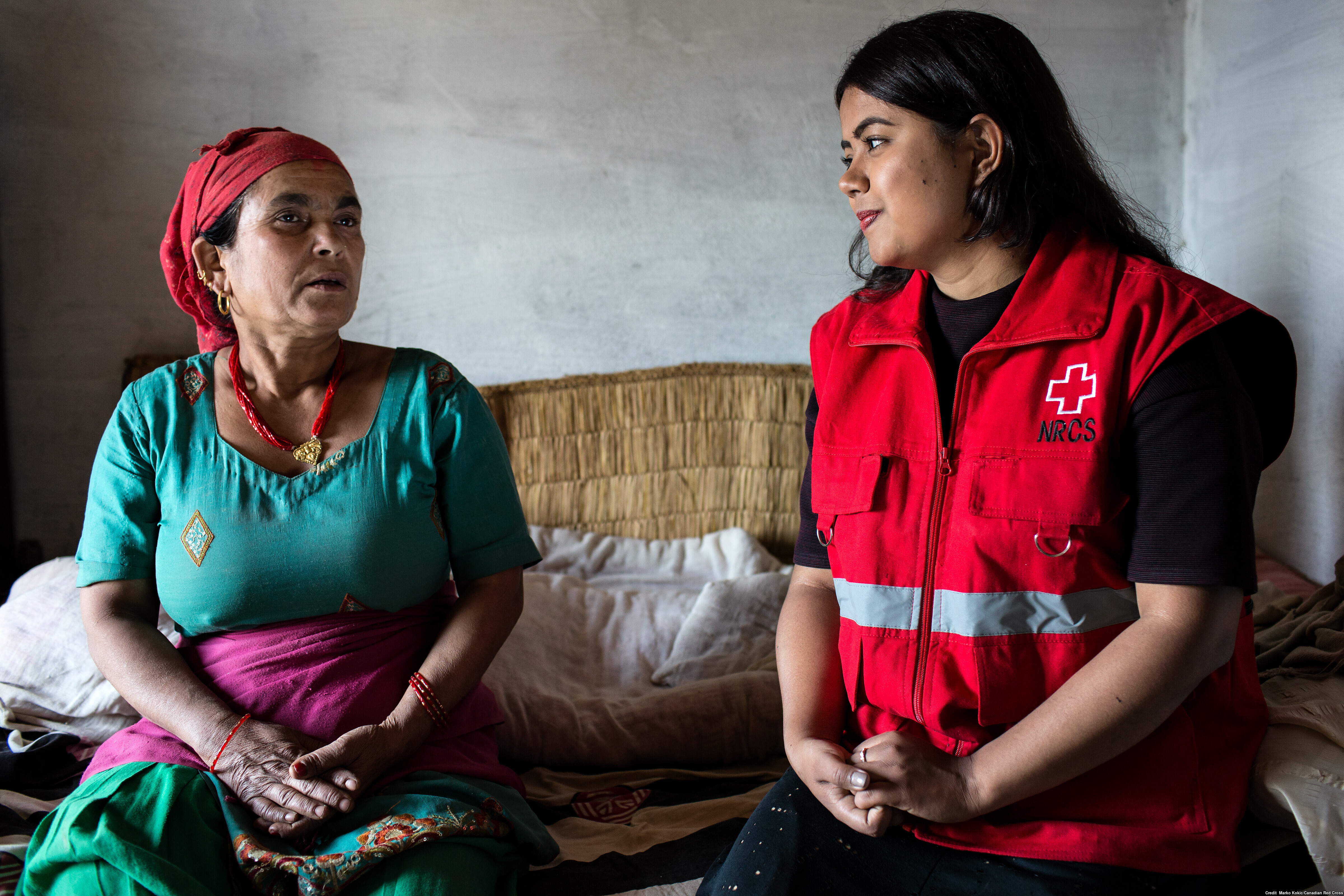 Une femme assise sur un lit discute avec une autre femme vêtue d’une veste rouge.