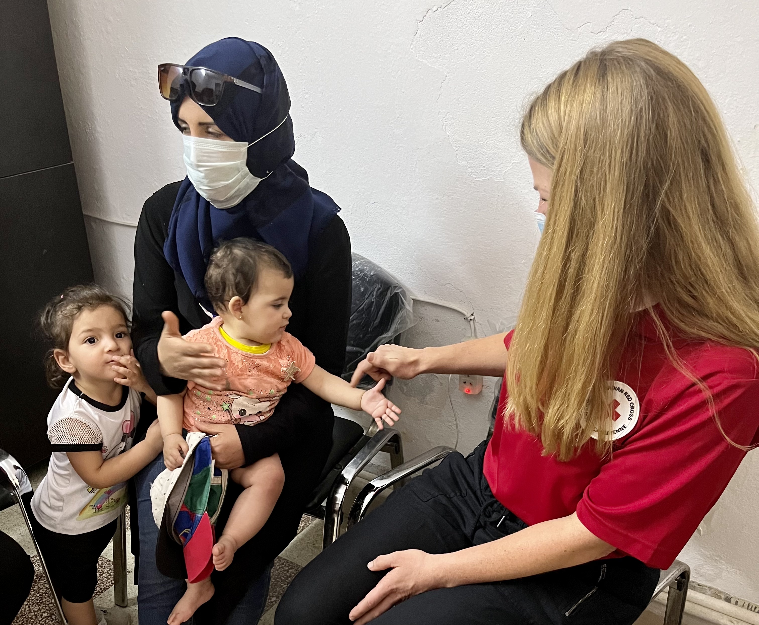 Une femme tenant un bébé est assise entre un enfant et une femme vêtue d’un t-shirt de la Croix-Rouge.